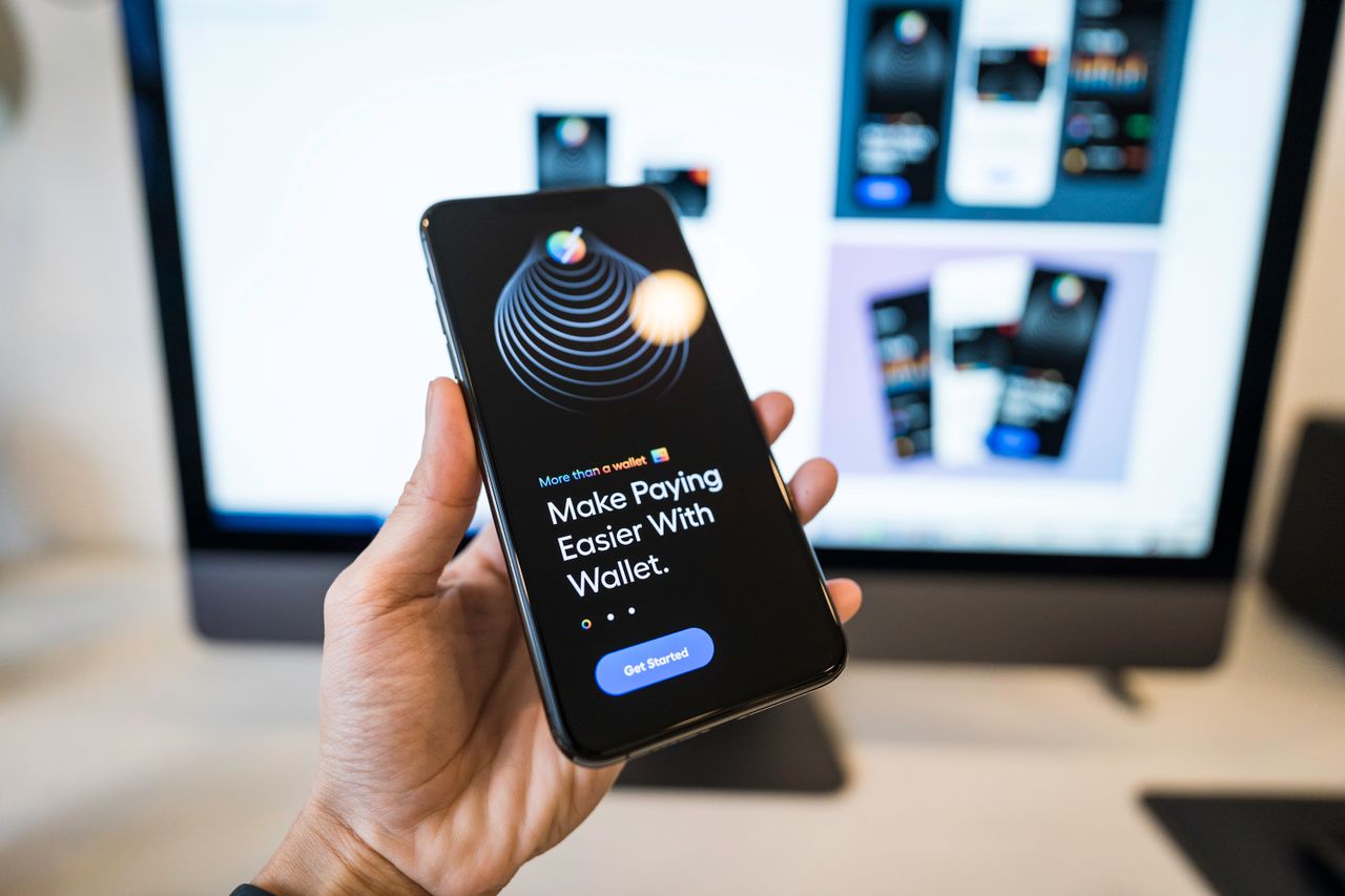 Main display of a smartphone in hand showing a digital wallet app interface, with a blurred monitor in the background.
